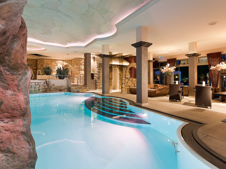 Innenpool mit beleuchteter Decke, stilvollen Säulen und gemütlichen Sitzbereichen und Wellness-Liegen im Hotel Der Heidehof